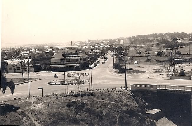 A black and white photograph of the area in the mid twentieth century, looking along a main road towards a roundabout, with a hotel, shopfronts and painted signage on the left and open ground and a bridge on the right.